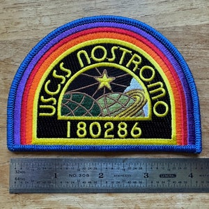 May include: A black and yellow embroidered patch with a rainbow border. The patch features the text "USCSS Nostromo" and the number "180286".