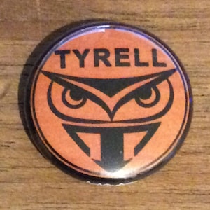 May include: An orange and black round button with the word "TYRELL" in white letters and an owl graphic with a black and white design.