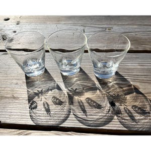 May include: Three clear, cone-shaped glasses with a wheat design etched into the bottom. The glasses are on a weathered wooden surface, casting shadows that mirror the wheat pattern. The glasses are empty.
