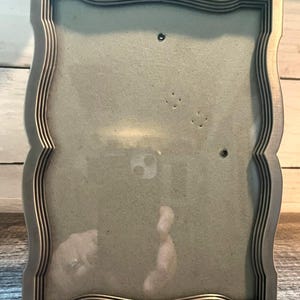 May include: A silver-toned picture frame with a wavy, scalloped edge design. The frame features a multi-layered, stepped border profile and a clear glass front panel. The interior of the frame contains a plain, light gray backing board.