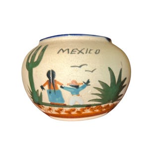 May include: A small, round ceramic vase with a cream-colored base and a blue rim. The word "MEXICO" is written across the top. The vase features a painted scene of two figures, birds, and cacti. The base has a brown and orange border.