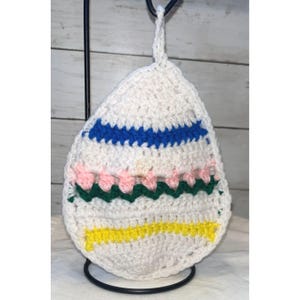 May include: White crochet Easter egg-shaped hanging decoration with blue, pink, green, and yellow stripes. The egg is suspended from a black metal stand. The crochet design features a loop for hanging.