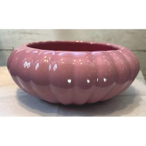 Op de afbeelding: Een glanzende, roze keramische kom met een pompoenachtig ontwerp. De kom heeft een brede, open bovenkant en een geribbelde buitenkant. De binnenkant is een donkerdere tint roze. Deze decoratieve kom is geschikt voor woondecoratie.