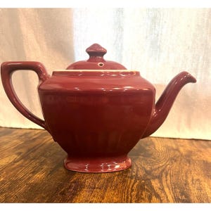 Op de afbeelding: Een bordeauxrode keramische theepot met bijpassend deksel. De theepot heeft een gebogen tuit, een grote handgreep en een kleine knop op het deksel. De theepot is effen bordeauxrood van kleur en lijkt in goede staat te verkeren.