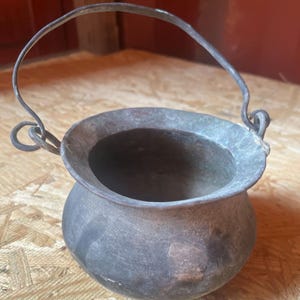 May include: A small, round, dark gray metal cauldron with a wide, flared rim and a thin, arched wire handle attached to two side loops. The surface of the vessel has a weathered, matte texture with subtle variations in color and tone.