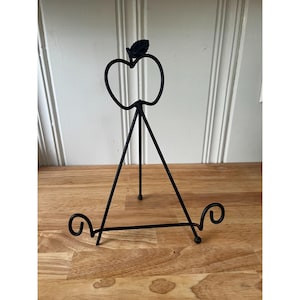 May include: Black metal tabletop display stand in the shape of an apple. The stand has a three-legged base and decorative swirls at the bottom. The apple design includes a leaf at the top.