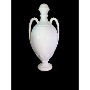 May include: A white ceramic amphora-shaped vase with a lid, two handles, and a pedestal base. The vase features decorative leaf and ribbed detailing. The overall design is symmetrical and elegant, suitable for home decor.