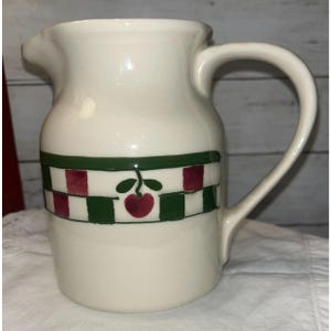 Puede incluir: Una jarra de cerámica de color crema con una banda decorativa alrededor del centro. La banda consiste en un patrón de cuadros verdes y granates intercalado con una pequeña ilustración de una cereza roja con hojas verdes en el frente.