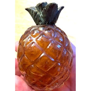 May include: A vintage amber-colored glass pineapple-shaped bottle with a detailed, textured surface. The top features a dark, metallic-looking pineapple crown. The bottle is held in a hand, showcasing its size and design.