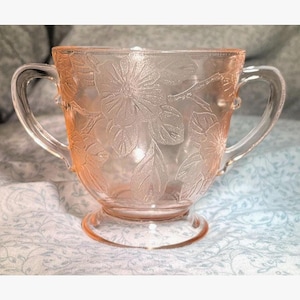 Puede incluir: Un azucarero vintage de cristal rosa con dos asas y una base de pedestal. El cuenco está decorado con un motivo floral en relieve de flores y hojas. El cristal tiene una calidad translúcida.