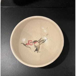 May include: Off-white ceramic bowl with a hand-painted hummingbird design. The hummingbird is depicted in flight, with red, black, and white details, and is feeding from a red flower. The bowl has a simple, rounded shape.