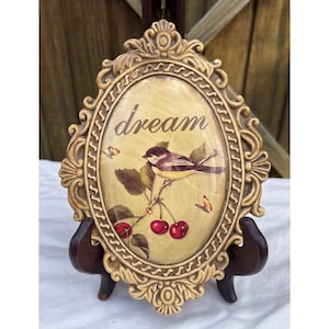 May include: An oval-shaped decorative plaque with a cream-colored frame. The plaque features a bird perched on a branch with cherries, butterflies, and the word "dream". The frame has ornate detailing and is displayed on a wooden stand.