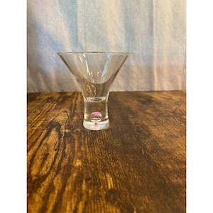 May include: A clear, stemmed martini glass with a flared bowl and a small, pink decorative element at the base. The glass sits on a dark wooden surface.