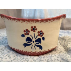 Könnte beinhalten: Cremefarbene Keramikschale mit rotem Rand und Boden. Die Schale hat ein blaues Schleifen- und Blumendesign in Rot und Blau. Sie hat eine geschwungene Form mit zwei kleinen Griffen.