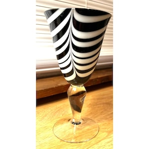 May include: A tall, elegant wine glass with a clear stem and base. The bowl features a striking black and white striped design. The glass is set on a wooden surface near a window.