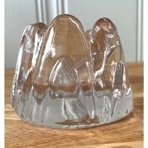 May include: A clear glass candle holder with a textured, mountain-like silhouette. The piece features a series of rounded, peak-shaped ridges that create a wavy, uneven top edge. The glass is transparent with subtle amber-toned highlights.