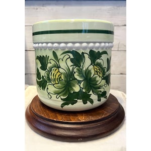 May include: A light green ceramic planter with a floral design in shades of green and yellow. The planter has a white beaded trim and sits on a dark brown wooden stand. The floral design is hand-painted.
