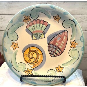 May include: A round ceramic plate with a coastal theme. The plate features hand-painted seashells, starfish, and blue and green accents. The plate is displayed on a black metal stand. The plate's diameter is approximately 12 inches.