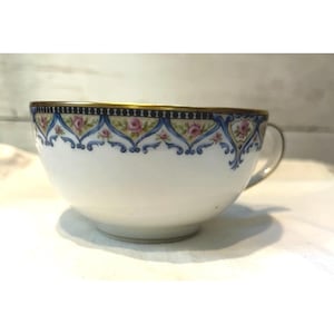 May include: White teacup with a gold rim and handle. The cup features a decorative band of pink roses, blue ribbons, and gold accents. The teacup is a classic design.