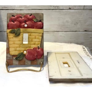 以下が含まれることがあります： 赤いリンゴと緑の葉が入ったバスケットが描かれた装飾的なライトスイッチカバー。 バスケットは黄色と茶色で、背景はチェック柄です。 カバーは金色の金属製スタンドに展示されています。