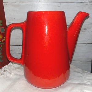 May include: A vibrant red ceramic watering can with a curved handle and a long spout. The watering can has a glossy finish and a rounded body. The background is a white surface and a wooden wall.