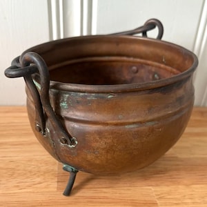 May include: A round copper pot with a dark, weathered patina and visible green oxidation. The pot features three short metal legs, a wide open top, and two curved metal handles attached to the sides with rivets.