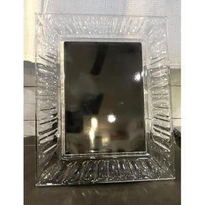 Puede incluir: Un marco de fotos rectangular hecho de vidrio transparente con un borde grueso y texturizado que presenta crestas verticales repetitivas. El centro del marco es una superficie lisa, oscura y reflectante que contrasta con los bordes.