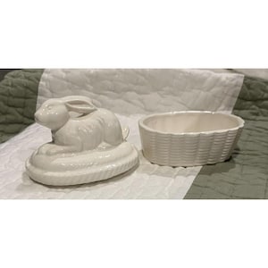 May include: A white ceramic Easter bunny figurine with a basket-weave style dish. The bunny is lying down on a decorative base. The dish is oval-shaped and has a basket-weave pattern. The set is likely for Easter.