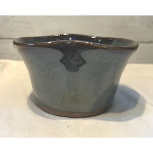 May include: A handmade ceramic bowl with a wavy rim. The bowl has a gray-blue glaze with a brown rim and base. The pottery bowl is suitable for serving food or as a decorative piece.