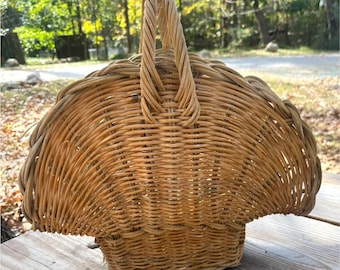 May include: A woven wicker basket with a handle. The basket is light brown and has a rounded shape with a scalloped edge. The handle is made of twisted wicker and is attached to the top of the basket.