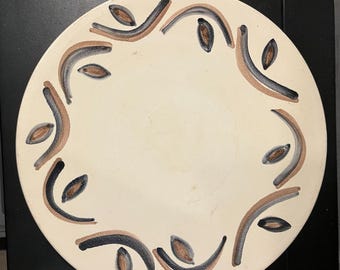 May include: A round, off-white ceramic plate with a decorative border. The border features a repeating pattern of brown, gray, and black curved lines and leaf-like shapes. The plate is set against a black surface.