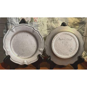 Può includere: Due piatti in ceramica grigia con bordi smerlati, esposti su supporti neri. Un piatto è rivolto verso l'alto, l'altro verso il basso, rivelando il testo "Conchitos Colonial by Costa" e un simbolo nautico. I piatti hanno un aspetto rustico e strutturato.