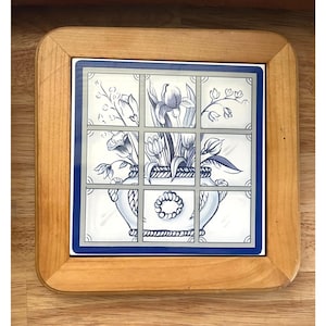 Puede incluir: Posavasos cuadrado de madera con una inserción de baldosa cerámica. La baldosa presenta un diseño floral azul y blanco de un jarrón con flores, enmarcado por un borde azul y un patrón de cuadrícula. El posavasos tiene un marco de madera marrón claro.