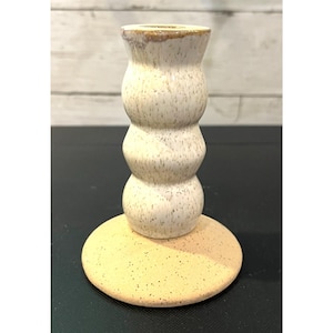 May include: A handmade ceramic candlestick holder with a speckled, off-white finish. The candlestick has a stacked, rounded design and sits on a round, speckled base. The top rim has a brown glaze.