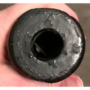 Op de afbeelding: Een close-up van een zwart, cirkelvormig object met een centraal gat. Het object heeft een getextureerd, verweerd uiterlijk, met zichtbare onvolkomenheden op het oppervlak. Het gat lijkt donker en diep, in contrast met de lichtere randen van het object.