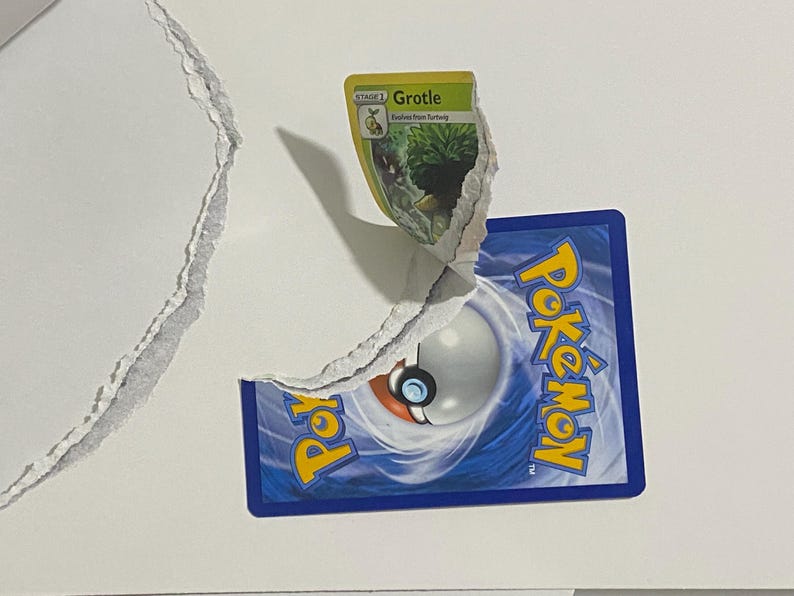 May include: A torn white surface reveals a Pok&eacute;mon trading card. The card's back is blue with the Pok&eacute;mon logo and a Pok&eacute; Ball design. A Grotle card is partially visible, with green and yellow accents.