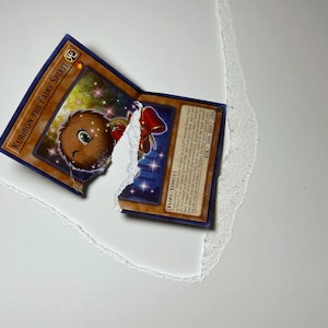 May include: A torn Yu-Gi-Oh! trading card featuring "Kuriboh the Fairy Spirit." The card has a brown and blue border with a cartoon illustration of a brown creature with a red bow. The card is partially torn, revealing a white background.