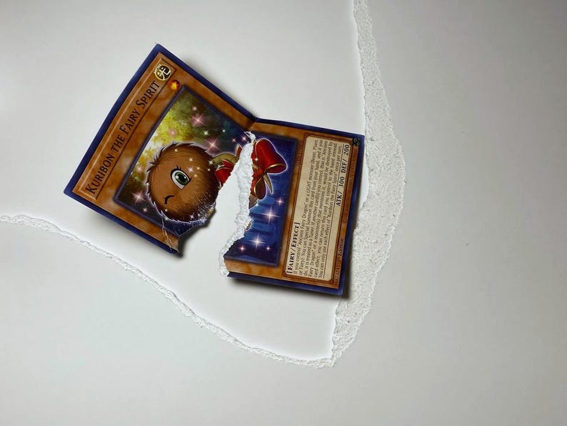 May include: A torn Yu-Gi-Oh! trading card featuring "Kuriboh the Fairy Spirit." The card has a brown and blue border with a cartoon illustration of a brown creature with a red bow. The card is partially torn, revealing a white background.