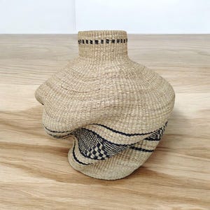 May include: A woven basket with a unique, asymmetrical shape. The basket is made of natural-colored fibers with a dark blue and black geometric pattern around the base. The rim of the basket has a black band.