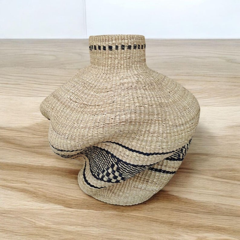 May include: A woven basket with a unique, asymmetrical shape. The basket is made of natural-colored fibers with a dark blue and black geometric pattern around the base. The rim of the basket has a black band.