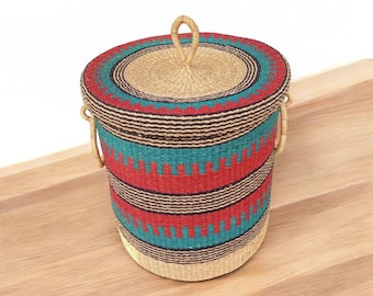 May include: A cylindrical woven storage basket with a lid. The basket features a vibrant pattern of red, teal, and black horizontal stripes. The lid has a woven handle. The basket is sitting on a wooden surface.