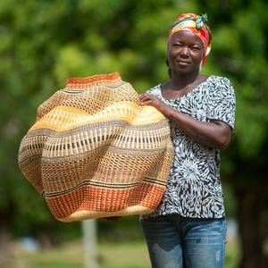 May include: A large, handwoven basket with a unique shape and a blend of natural and orange hues. The basket is held by a person wearing a black and white patterned top and blue jeans. The basket's design features intricate weaving patterns.