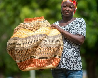 May include: A large, handwoven basket with a distinctive shape, in natural and orange tones. The basket is held by a person wearing a black and white patterned top and blue jeans. The basket's design showcases intricate weaving.