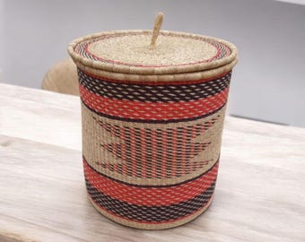 May include: A cylindrical woven basket with a lid. The basket features alternating bands of red and dark brown, with a central design. The lid has a small handle. The basket is made of natural fibers.