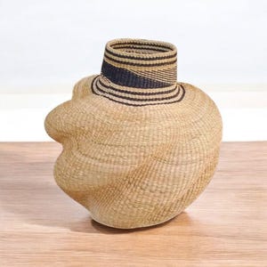 May include: A handwoven basket with an unusual, organic shape. The basket is made of natural-coloured fibres, with a cylindrical neck featuring black and tan horizontal stripes. The body of the basket has a rounded, asymmetrical form.