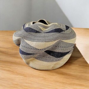 May include: A woven basket with a unique, organic shape. The basket features a pattern of alternating beige, gray, and navy blue horizontal stripes. The basket is sitting on a wooden surface.