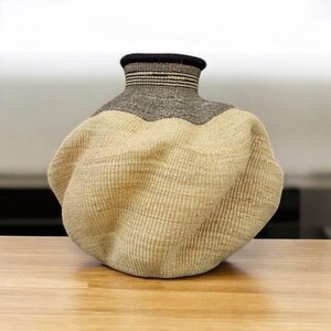 May include: A woven vase with a unique shape, featuring a beige, gray, and black color scheme. The vase has a textured surface and a wide, rounded body that tapers to a narrower neck. The top is black with horizontal stripes.