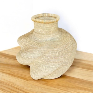 May include: A woven basket with a unique, asymmetrical shape. The basket is made of natural-colored fibers, with a wide, rounded body and a narrower, cylindrical neck. The basket sits on a wooden surface.