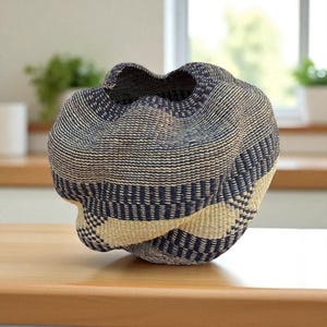 Può includere: Un cesto intrecciato con una forma unica e organica. Presenta una combinazione di fibre blu navy e beige naturale, creando un motivo a righe. La parte superiore aperta ha un bordo ondulato, che aggiunge al suo design distintivo.