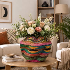 May include: A colorful woven basket vase filled with an arrangement of flowers. The vase has a vibrant pattern of pink, green, orange, and black stripes. The flowers include white and pink roses, and greenery. The vase sits on a round wooden coffee table.